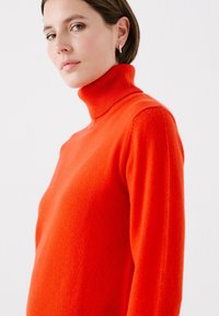 Maglione arancione a collo alto, realizzato in materiale a maglia morbida, con una silhouette aderente e un colletto a coste, caratterizzato da una superficie liscia e senza motivi visibili.