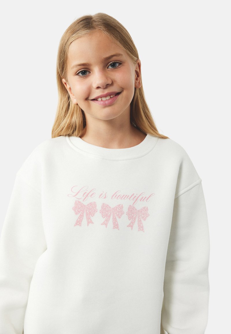Witte sweatshirt met drie roze strikgraphics en de tekst "Het leven is mooi" in een cursieve lettertype. Zachte stof, ruime pasvorm, ronde hals.