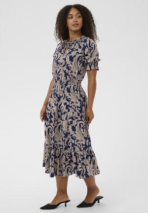 Laivastonsininen mekko, jossa on kermaan vivahtava paisley-kuvio, lyhyet puffihihat, istuva vyötärö ja levenevä helma. Päällä mustat korkokengät ilman kantaa.