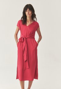 Donna con capelli lunghi e scuri, che indossa un vestito rosso midi con maniche corte, una cintura annodata in vita e fessure laterali, in piedi con le mani nelle tasche.