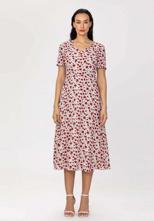 Femme portant une robe midi blanche à imprimé floral rouge, manches courtes et col en V, associée à des talons blancs à brides à la cheville, debout devant un fond uni.