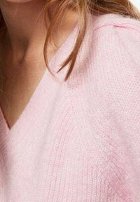 Svetle růžový pletený svetr s výstřihem do V, který ukazuje jemnou texturu a viditelný žebrový vzor. Detailní záběr na tkaninu oděvu.