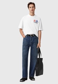 Camiseta blanca de manga corta con un gráfico colorido impreso, combinada con pantalones vaqueros sueltos de color azul oscuro y zapatos negros. Una bolsa tote negra completa el look.