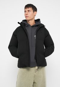 Polstret svart jakke med hette, glidelås og to frontlommer, brukt over en mørkegrå hettegenser. Glatt tekstur, minimalistisk design.