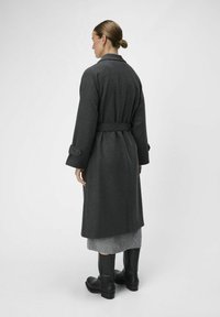 Manteau long gris en tissu texturé, doté d'une taille ceinturée, de poignets boutonnés et d'une silhouette droite, porté sur une jupe grise et des bottes noires.