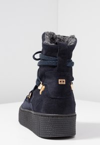 Námořnické modré semišové kotníkové boty s šedou kožešinou uvnitř, zaoblenou špičkou, silnou strukturovanou černou podrážkou a zlatými akcenty na stranách.