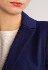 Granatowa marynarka z wyraźnymi klapami i gładkim materiałem, zestawiona z teksturowaną jasnobeżową bluzką o subtelnym prążkowaniu.