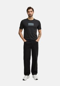 Camiseta de algodón negra con un logo de Calvin Klein en degradado azul en el centro, combinada con pantalones negros y zapatillas deportivas blancas.