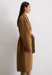 Femme aux longs cheveux noirs portant un manteau en laine marron ceinturé, se tenant de profil devant un mur clair et uni.