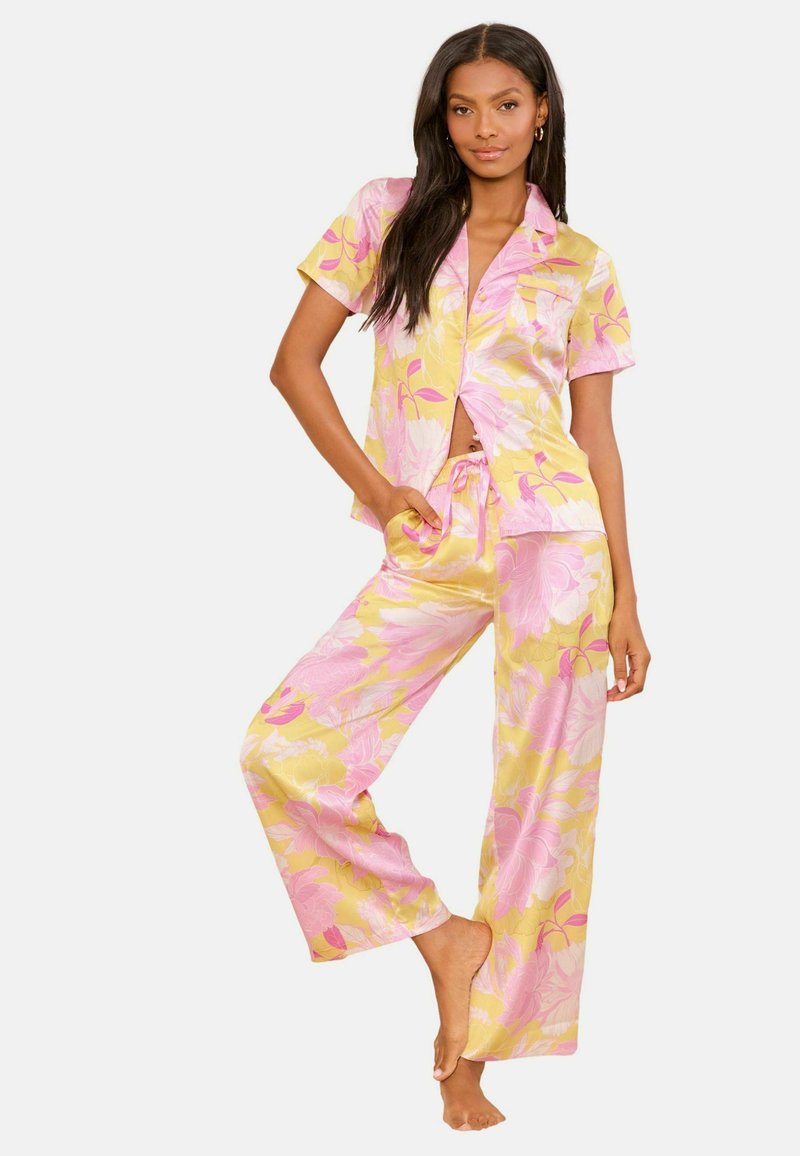 Vrouw staat op blote voeten in een satijnen pyjama-set met gele en roze bloemen, bestaande uit een kortärmelig overhemd met knopen en broek met wijd uitlopende pijpen, hand in de zak.