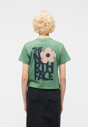 Άτομο με κυματιστά ξανθά μαλλιά, φανερό από πίσω, φοράει πράσινο κοντό μπλουζάκι με το κείμενο "The North Face" και ένα ροζ γραφικό λουλούδι.