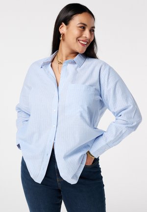 Vrouw die glimlacht, gekleed in een lichtblauw-wit gestreept overhemd met knopen en een zak, donkere spijkerbroek, gouden sieraden, poserend met handen in de zakken.