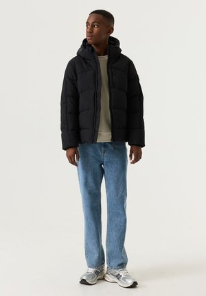 Teenager steht nach links gewandt, trägt eine schwarze Steppjacke, ein hellgraues Shirt, blaue Jeans und weiße Turnschuhe vor schlichtem Hintergrund.