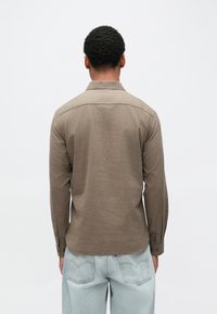 Hnedá, dlhý rukáv košeľa z texturovanej látky s golierom na gombíky, pohľad zozadu, v kombinácii so svetlomodrými džínami.