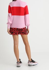 Femme portant un pull rayé rose et rouge, une mini-jupe à motifs rouges et noirs, et des baskets roses et noires, debout devant un fond blanc.