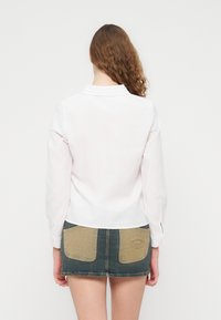 Λευκό πουκάμισο με μακριά μανίκια και γιακά, με ίσια γραμμή. Συνδυάζεται με φούστα από τζιν, με δύο ταμπά τσέπες ως διακοσμητικό στοιχείο.