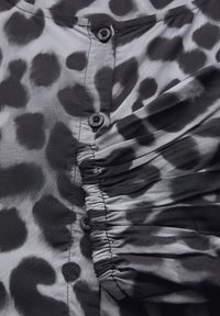 Blouse à motifs noirs et gris avec une texture lisse, dotée de boutons, de détails de tissu froncé et d'un design de col décontracté.