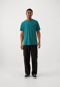 Camiseta turquesa de manga corta, combinada con pantalones negros y zapatillas negras. Persona lleva gafas de sol con lentes tintadas doradas. Diseño sencillo, ajuste relajado.