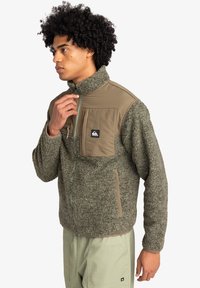 Fleecejacka i olivgrön, med en strukturerad kropp, slät panel fram, hög krage, dragkedjestängning och bröstficka.