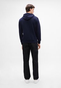 Granatowy sweter z kapturem z dopasowanym dołem i ściągaczem, zestawiony z ciemnoniebieskimi dżinsami i białymi sneakersami, widok od tyłu.