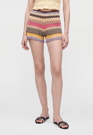 Person iført flerfarvede zigzag-mønstrede strikkede shorts, beige rynket crop top og sorte slidesandaler med gulddetaljer på en ensfarvet baggrund.