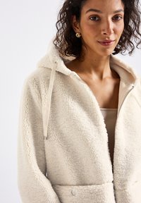 Femme portant un hoodie zippé en sherpa couleur crème avec capuche et cordons, débardeur beige en dessous, et boucles d'oreilles dorées.