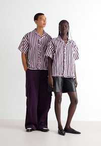Duas modelos estão juntas. Elas usam camisetas listradas de mangas curtas em roxo e branco, combinadas com calças largas roxas e calções de couro pretos.