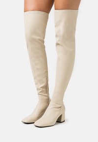 Botas beige hasta la rodilla, hechas de material liso, con punta cuadrada, tacón grueso y cremallera lateral para facilitar su uso.