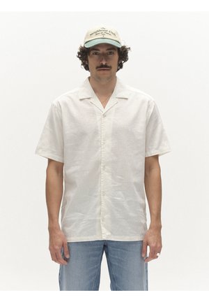 Hombre con bigote que lleva una gorra de béisbol beige, una camisa blanca de manga corta abotonada y vaqueros azul claro, de pie frente a un fondo blanco.