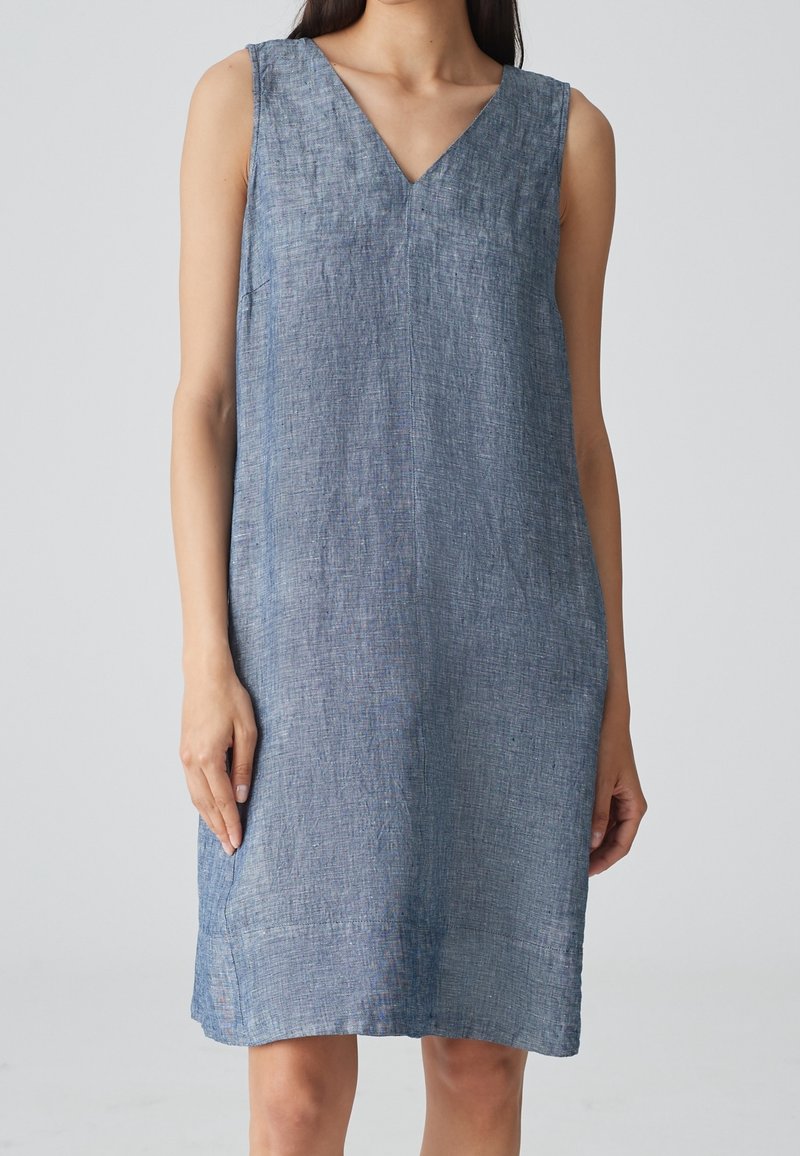 Femme portant une robe sans manches texturée bleu clair, longueur genou, avec un col en V, debout devant un fond uni gris clair.