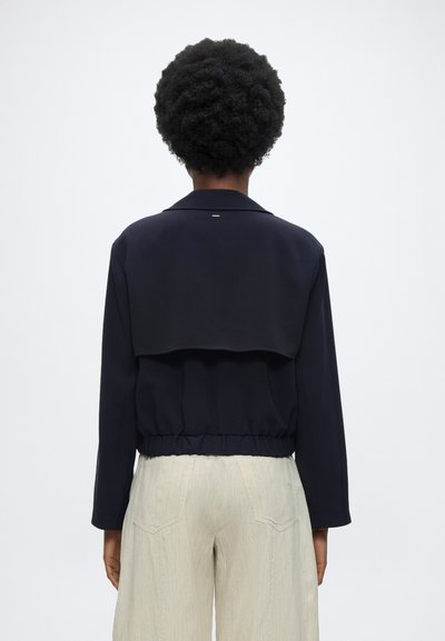 Personne aux cheveux courts et bouclés porte une veste noire courte et un pantalon beige clair, vue de dos sur fond blanc uni.