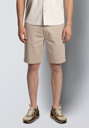 Mann iført beige shorts, lys beige knappeskjorte og beige og mørkebrune joggesko, stående mot en ensfarget grå bakgrunn.