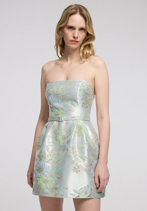 Mujer con vestido sin tirantes hasta la rodilla con patrón floral en azul claro, verde y dorado y un cinturón fino, de pie contra un fondo liso.
