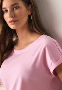 Tricou roz deschis cu mâneci scurte și decolteu rotund, fabricat din material moale. Buzele sunt într-o nuanță neutră, iar cerceii cu cerc auriu evidențiază aspectul.