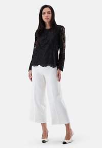 Schwarzes Spitzenoberteil mit langen Ärmeln und Blumenmuster, mit gewelltem Saum; kombiniert mit weißen, weit geschnittenen Hosen und zweifarbigen Absätzen mit schwarzen Spitzen.