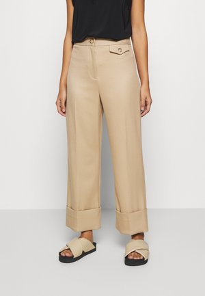 Kobieta ubrana w beżowe spodnie o szerokich nogawkach z podwiniętymi mankietami, czarną bezrękawnikową bluzkę i beżowe sandały na paski z czarną podeszwą.