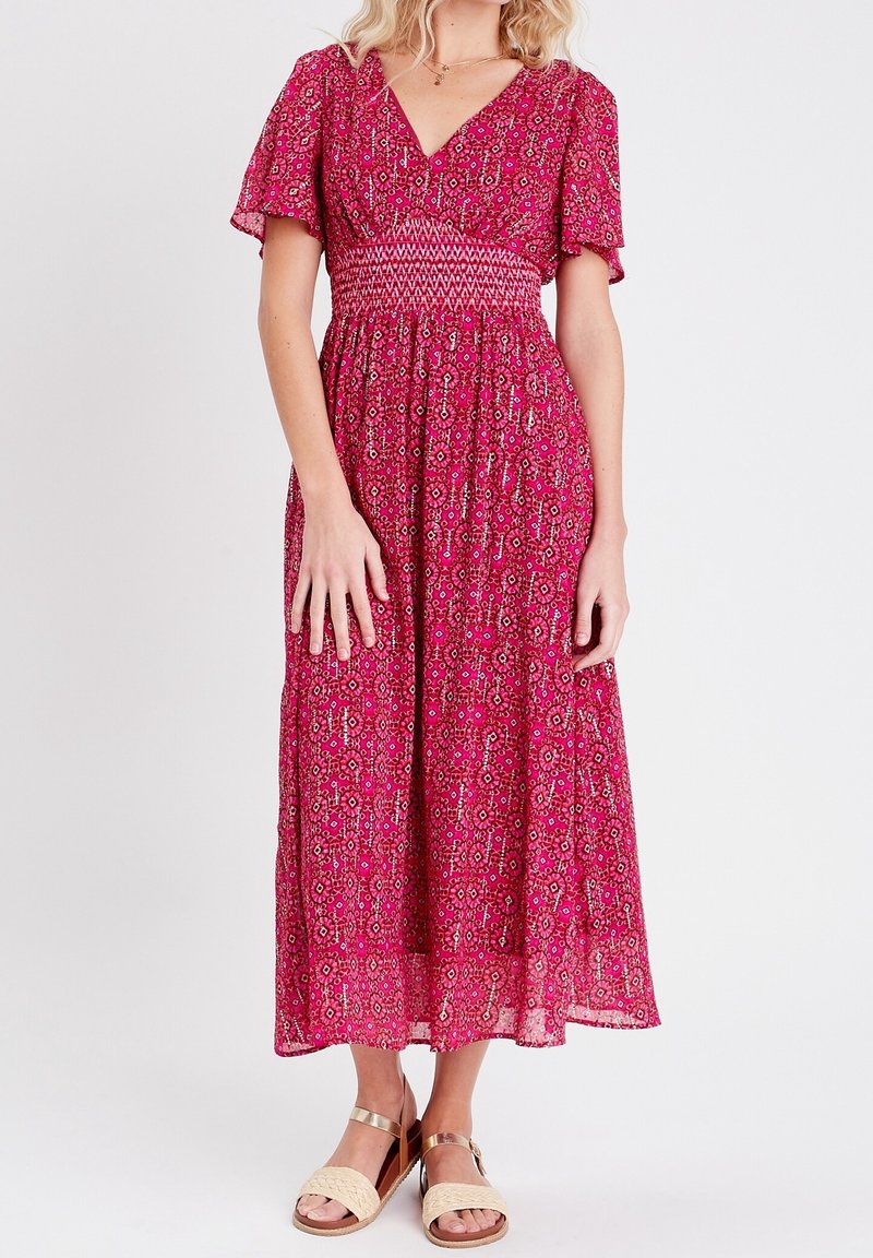Robe maxi florale rose avec des manches courtes et un décolleté en V. Possède une taille smockée et une jupe fluide. Associée à des sandales beiges à lanières.