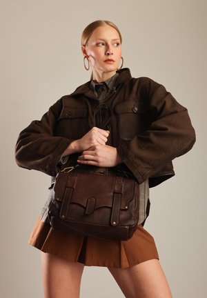 Hnědá kožená kabelka s klopou a dvěma předními kapsami, se zlatými akcenty a detailem šití, kterou drží modelka v hnědém saku.