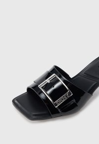 Μαύρο σανδάλι με λουρί από πατέν τα και τετράγωνη μύτη. Διαθέτει φαρδύ λουρί με μεταλλική αγκράφα και ανάγλυφο λογότυπο. Σχεδίαση με επίπεδη σόλα.