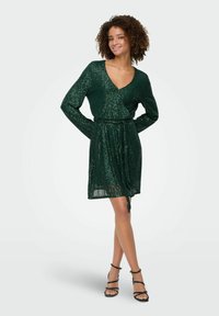 Zelena obleka s sequini, z dolgimi rokavi, v-vratom in nastavljivim pasom, ki ima prosojno teksturo in tekočo silhueto.