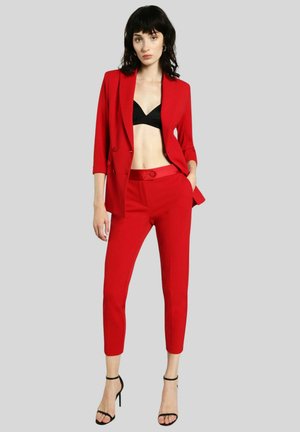 Donna con capelli neri indossa un blazer rosso sartoriale e pantaloni, reggiseno nero e sandali neri con tacco alto, in piedi davanti a uno sfondo semplice.