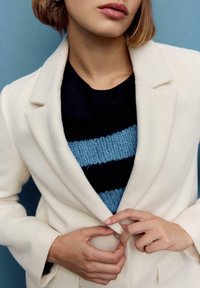 Cappotto bianco di lana sopra un maglione a righe blu navy e azzurro. Il cappotto ha un collo a rever e due tasche frontali, con una texture liscia.