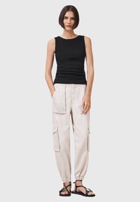 Top senza maniche nero, pantaloni cargo beige con multiple tasche, polsini elasticizzati e sandali neri con cinturini, in piedi su uno sfondo neutro.