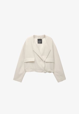 Lyhennetty vaaleanbeige bleiseri, jossa on kauluksessa sakara, kaksi etutaskua ja pyöreä solki-detail sivussa. Tasainen kangas, jossa on minimaalinen tekstuuri.