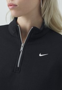 Černá mikina na zip vyrobená z měkké látky s vysokým límcem, se stříbrným zipem a malým bílým logem Nike na prsou.