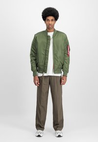Olive green bomber jacket with a zip front, ribbed cuffs, and side pockets. White T-shirt underneath and brown loose-fitting trousers.