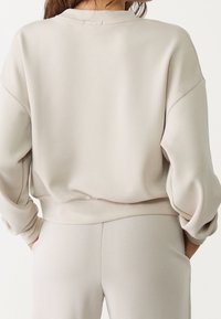 Jasno beżowy sweatshirt o luźnym kroju, z szerokimi rękawami i ściągaczem u dołu. Gładka tekstura, okrągły dekolt i minimalne detale szwów widoczne.