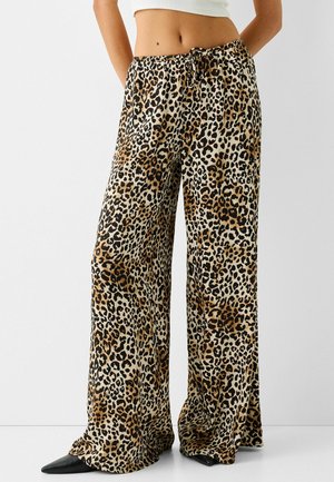 Mujer con pantalones de pierna ancha con estampado de leopardo, cintura con cordón y zapatos negros puntiagudos, de pie contra un fondo liso.