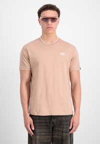 Svetloružové tričko s krátkym rukávom a okrúhlym výstrihom obsahuje malé biele logo. Model má na sebe šedé nohavice a svetlé slnečné okuliare.