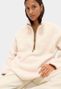 Polarowy pulower w kolorze kremowym, z pół-zamkiem, jasnobrązowym skórzanym uchwytem, miękką teksturą i szerokimi rękawami, noszony na jasnych spodniach.